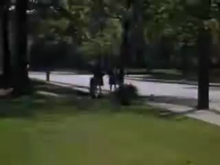 The still from 'Connie and Shirley reel #1 - 1941' captures a sunny day in a suburban setting. In the foreground, lush green grass contrasts with the paved road running through the scene. Silhouetted figures are seen walking along the street, surrounded by trees that provide dappled shade. The overall ambiance conveys a leisurely and carefree moment from the early 1940s.