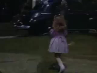 The still features a young girl in a light purple dress, running playfully on a grassy area. She has long, wavy hair and appears to be enjoying herself in an outdoor setting, where a vintage car is partially visible in the background. The scene, captured on Kodachrome film from 1941, conveys a sense of nostalgia and innocence typical of childhood moments from that era.