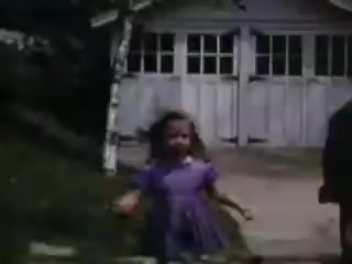 The still shows a young girl in a purple dress running joyfully outside a building, likely a garage or shed. The scene is vibrant, typical of Kodachrome's rich color palette, with a leafy tree nearby and a sense of playful nostalgia from the early 1940s. The background suggests a suburban or rural setting.