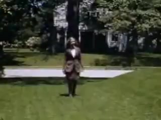The 16mm still from 'Connie and Shirley reel #1 - 1941' features a figure walking across a lush green lawn, dressed in formal attire. In the background, a house can be seen among trees, capturing a serene, domestic scene typical of the early 1940s. The vibrant colors associated with Kodachrome film highlight the outdoor setting and the details in the clothing.
