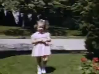 The still captures a young girl dressed in a light pink dress, standing on a grassy area with greenery around her. She has her hands clasped and appears to be smiling, suggesting a joyful moment. The background includes a well-maintained garden with flowering plants, and a clear day adds a vibrant feel to the scene, characteristic of Kodachrome’s rich color reproduction in 1941.