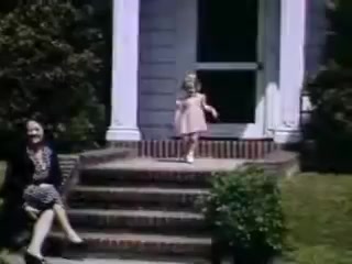 The still features a sunny outdoor scene from 1941, showcasing a young girl in a pink dress joyfully walking down the steps of a house. She exhibits a sense of playfulness and innocence. Seated on the steps is an older woman, likely a family member, in a patterned dress, appearing relaxed and enjoying the moment. The frame is vibrant, characteristic of Kodachrome film, with lush greenery surrounding the entrance.