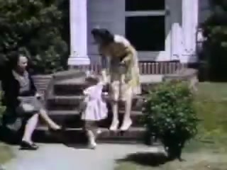 The still from 'Connie and Shirley reel #1 - 1941' depicts two women seated on a set of steps outside a house, with one woman leaning over to assist a small girl dressed in a light-colored dress. The scene captures a sunny day, with vibrant colors typical of Kodachrome film from that era, and a well-maintained lawn surrounding the home. The atmosphere appears warm and familial, suggesting a moment of play or interaction among the subjects.