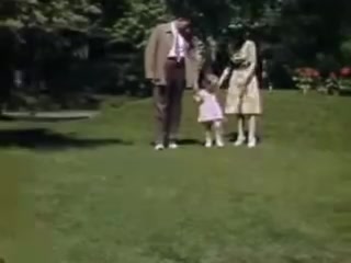 The still captures a moment in a vibrant garden setting. A man and a woman stand together, holding hands with a young girl dressed in a pink dress. The scene conveys a sense of family togetherness amidst lush greenery and blooming flowers, characteristic of leisure time in the early 1940s. The use of Kodachrome film enhances the vivid colors of their clothing and the natural surroundings.