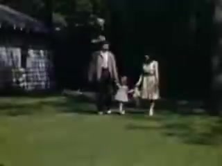 The still shows a family walking together on a green lawn. A man, possibly a father, and a woman, likely the mother, are holding hands with a young girl between them. They are dressed in mid-20th century attire, and there is a house or structure in the background. The scene is vibrant, reflecting the Kodachrome film's vivid colors typical of the era.