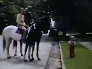 The still from 'Connie and Shirley reel #1 - 1941' features two riders on horseback, one on a black horse and the other on a white horse. They are positioned on a gravel path in a park-like setting, with trees in the background. In the foreground, a young child stands on the grass, watching the riders. The colors are vibrant, characteristic of Kodachrome film from that era.