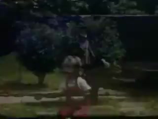 The still from 'Connie and Shirley reel #1 - 1941' features a vibrant outdoor scene, likely captured in a backyard or park setting. In the foreground, a child wearing a white shirt and red skirt is engaged with an adult in a playful manner. The background is filled with greenery and trees, typical of Kodachrome’s rich colors from that era, evoking a sense of nostalgia and warmth. The overall composition suggests a moment of joy and interaction between the figures.