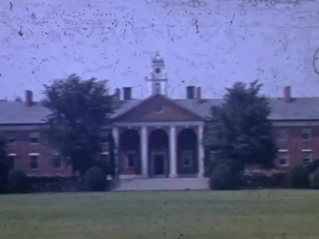 The still captures a prominent brick building set in a grassy area, likely a school or institutional structure, given its classical architectural features. It features a central portico with columns and a clock tower. Surrounding trees add greenery to the scene, indicative of a mid-20th century setting, with the Kodachrome color palette and slight film grain suggesting it's from the early 1940s.