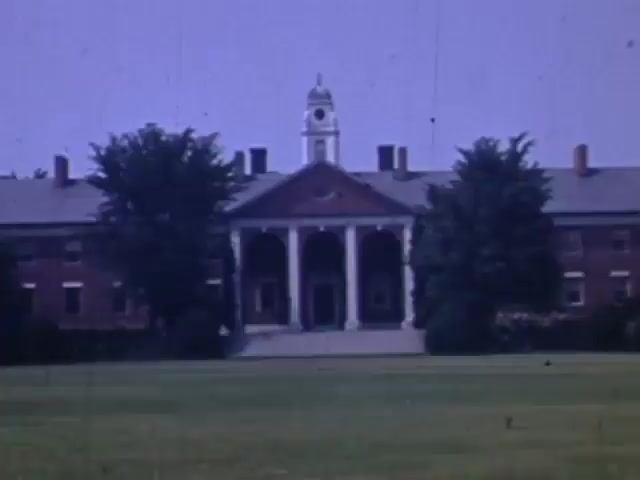 The still features a large brick building with a prominent clock tower and classical architecture, including a portico supported by columns. The surrounding area has well-maintained grass and trees, suggesting a formal educational or institutional setting. The image, captured in Kodachrome from 1941 to 1943, showcases vibrant colors typical of the film style, despite showing signs of age.