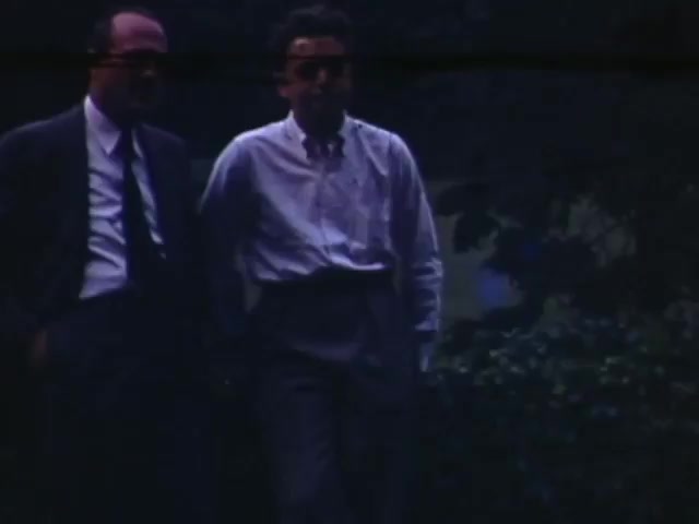 The still from 'Jack Reel 2' depicts two men casually walking side by side, likely outdoors. One man is dressed in a suit and tie, while the other wears a light-colored button-up shirt. The scene is characterized by soft natural lighting and a slightly blurred background, hinting at a relaxed moment in a park or garden setting. The colors have a vintage quality, typical of Kodachrome film from the early 1940s.