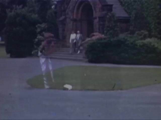 The still from 'Jack Reel 2' features a serene outdoor scene captured on Kodachrome, likely from the early 1940s. Two individuals, dressed in light-colored clothing, are seen walking down steps from a building with an ornate entrance, surrounded by well-maintained greenery and flowering plants. The overall composition conveys a peaceful, leisurely atmosphere typical of that era. The color quality reflects the vibrant hues characteristic of Kodachrome film, adding a nostalgic touch to the moment.