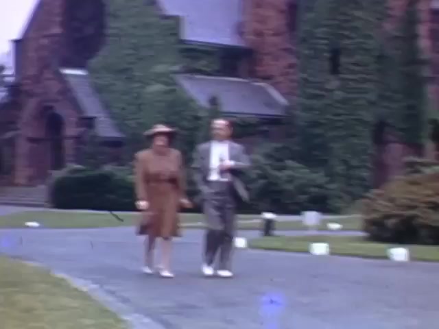 The still from 'Jack Reel 2,' captured in Kodachrome between 1941 and 1943, features a man and woman walking together on a pathway. The woman, dressed in a brown dress and hat, appears cheerful, while the man, in light gray attire, smiles as they stroll. The background includes ivy-covered buildings, enhancing the vintage charm of the scene. The colors are vibrant, characteristic of Kodachrome film, conveying a sense of leisure and companionship from that era.
