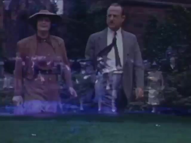 The still from 'Jack Reel 2,' shot on Kodachrome film between 1941 and 1943, depicts two figures walking in a garden setting. The person on the left is dressed in a tan outfit and hat, suggesting a casual or outdoor theme, while the figure on the right is in formal attire, indicating a contrast in style. The lush greenery and soft colors typical of Kodachrome enhance the nostalgic atmosphere, typical of early 20th-century leisure scenes.