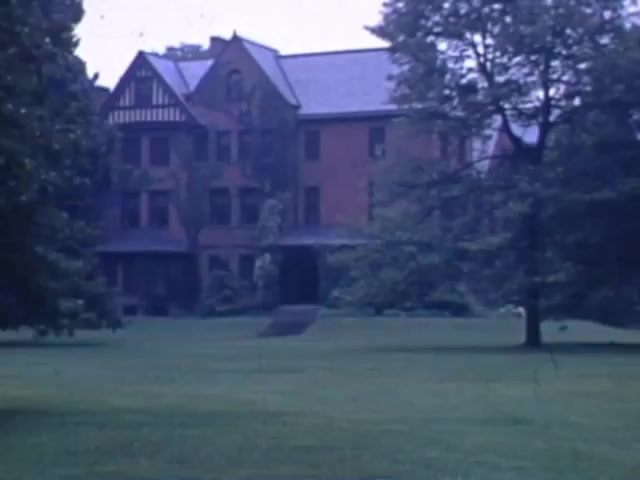 The still from 'Jack Reel 2' features a large, historic brick building set amidst a lush, green lawn. The architecture showcases elements typical of early 20th-century design, including multiple gables and large windows. The surrounding landscape includes a few trees, contributing to a serene, possibly rural atmosphere. The muted colors are characteristic of Kodachrome film, reflecting the period from 1941 to 1943.
