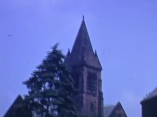 The still captures a church with a prominent tower, surrounded by lush greenery. The sky is a soft blue, indicative of a clear day. This image, part of a Kodachrome reel filmed between 1941 and 1943, likely reflects daily life or significant landmarks from that era. The colors are vibrant, typical of Kodachrome film, emphasizing the architectural details and natural surroundings.