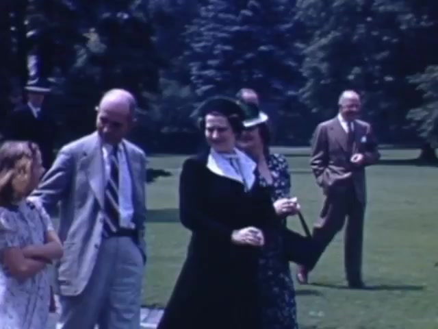The still features a casual outdoor gathering with several adults and a girl, likely in a park setting. The adults are dressed in formal or semi-formal attire typical of the early 1940s, with men in suits and women in dresses and hats. The background displays lush greenery, indicative of a leisurely day outdoors. The scene captures a moment of interaction among the group, suggesting a social or familial event during the early 1940s.