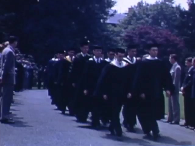 The still shows a group of young men in graduation caps and gowns, marching in a procession. They are likely graduates celebrating a significant academic milestone, possibly taking place outside in a park-like setting. The background features trees and a crowd of spectators lining the walkway, indicating a formal ceremony. The image captures the spirit of a celebratory moment from the early 1940s, presented in vibrant Kodachrome color.