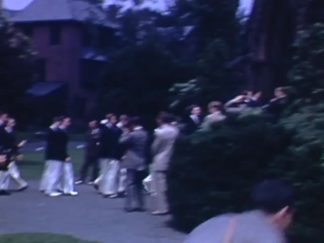 The still features a group of men in formal attire, likely from the early 1940s, gathered outdoors in a campus-like setting. Some are walking, while others appear to be conversing. The scene is framed by lush greenery and buildings in the background, with a relaxed atmosphere suggesting a social or academic event. The color palette reflects the typical Kodachrome vibrancy of that era.