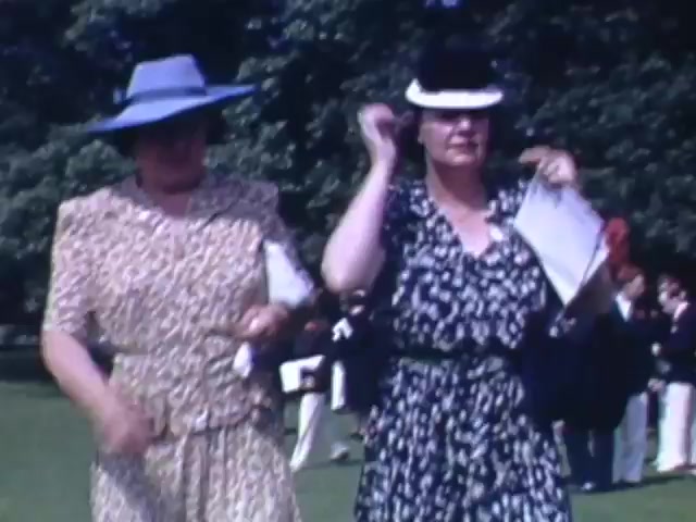 The still from 'Jack Reel 2,' shot on Kodachrome between 1941 and 1943, captures two women walking on a grassy area. The woman on the left wears a light blue hat and a leopard-print dress, while the woman on the right dons a black hat with a white band and a black dress with white spots. Both seem to be enjoying a social outing, with people visible in the background, suggesting a leisurely outdoor event typical of the era. The vibrant colors characteristic of Kodachrome enhance the scene's lively atmosphere.