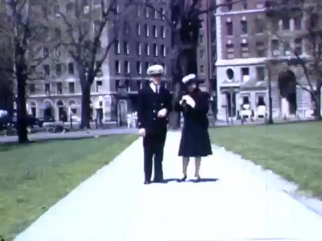The still depicts a scene from a park, likely in the early 1940s. A young man in a naval uniform and a woman in a stylish dress and hat walk together along a paved path. The background features a mix of trees and mid-century architecture, indicative of urban environments during that era. This moment captures a sense of camaraderie and leisure amidst the times of World War II. The Kodachrome color adds a nostalgic vibrancy typical of film from that period.