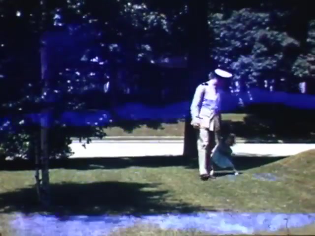 The still from 'Jack Reel 2' features a soldier in a uniform, possibly from the early 1940s, walking on a grassy area alongside a child. The background includes trees and a suburban street, suggesting a peaceful, domestic setting. The colors are vivid, characteristic of Kodachrome film from that era, indicating a moment of family life during World War II.