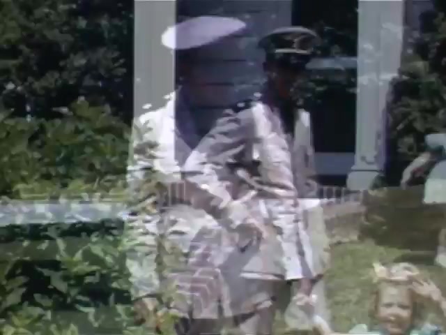 The still from 'Jack Reel 2' captures a moment likely from the early 1940s, featuring a man in a military uniform alongside a child and a woman. The Kodachrome colors are vibrant, highlighting greenery and the residential setting. The image features a blend of figures, indicating a possible double exposure effect, which adds a layer of complexity and nostalgia to the scene. The context suggests a familial gathering during wartime, reflecting the emotional themes of the era.
