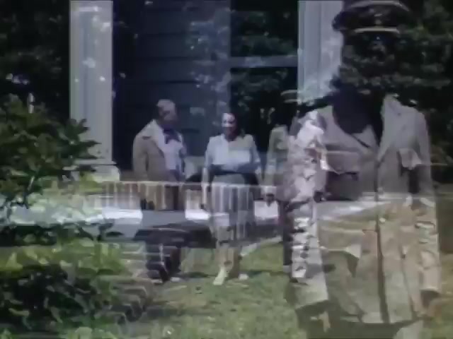 The still from 'Jack Reel 2' showcases a scene from the early 1940s, featuring a man and a woman, dressed in period-appropriate attire, engaged in conversation outdoors. The presence of a military figure in uniform hints at a possible wartime context. The vibrant Kodachrome colors capture the lush greenery surrounding them, suggesting a sunny day. The historical significance and personal interactions reflect a blend of domestic life and the impact of World War II during this era.