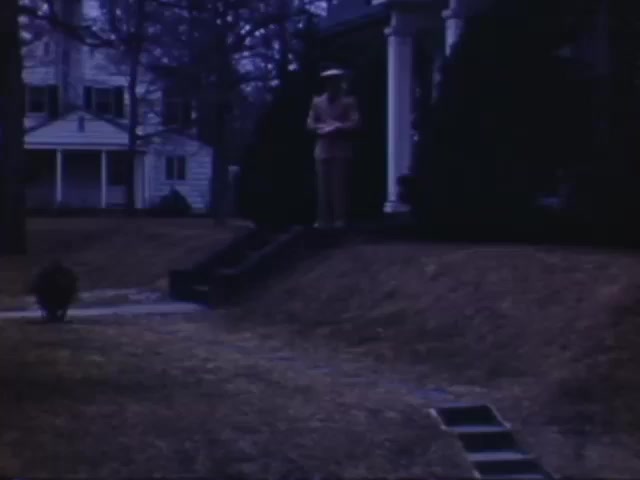 The still from 'Jack Reel 2' features a man dressed in a light-colored suit standing on the steps of a house. The scene captures a quiet suburban setting, with neatly maintained homes and sparse vegetation, indicative of early spring. The color cast suggests the typical Kodachrome richness, though it appears slightly muted, possibly due to aging or lighting conditions. The time frame spans from 1941 to 1943, hinting at a historical context reflective of that era.