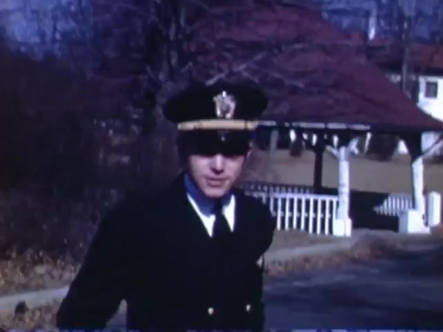 The still from 'Jack Reel 2,' captured on Kodachrome film, features a uniformed officer standing near a picturesque gazebo in a setting that reflects early 1940s aesthetics. The image is likely set in a suburban area, with trees in the background and a clear blue sky. The scene conveys a sense of authority and community typical of the era, likely filmed between 1941 and 1943.