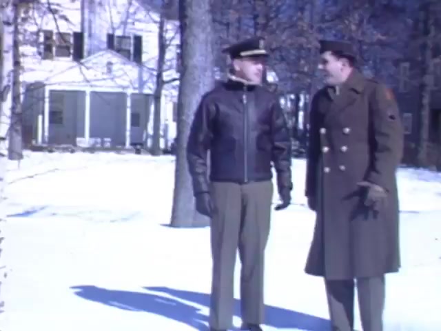 The still depicts two men conversing in a snowy outdoor setting, likely during the winter months of the early 1940s. One man is dressed in a leather flight jacket and cap, suggesting he may be a pilot or military officer, while the other is in a double-breasted military coat. In the background, there are trees and a house, indicating a suburban environment. The use of Kodachrome film suggests vibrant colors typical of the era. The timeframe likely reflects a moment of camaraderie or discussion related to military service, set against a picturesque winter backdrop.