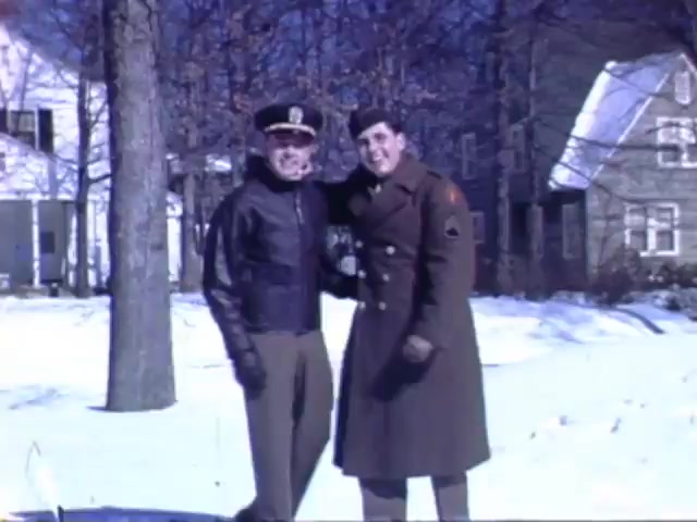 The still features two soldiers posing together in a snowy outdoor setting, likely in a suburban area. One is dressed in a leather flight jacket and military cap, while the other wears a long coat with military insignia. The backdrop includes residential houses, hinting at a home front scene during World War II. The Kodachrome colors are vivid, capturing the essence of the early 1940s.