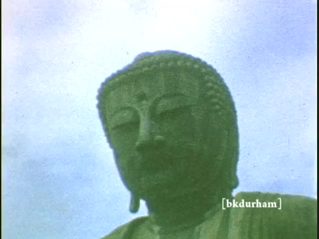 The still features a close-up of the Great Buddha (Daibutsu) in Kamakura, Japan, captured from an 8mm home movie likely shot between 1959 and 1960. The image captures the serene expression and intricate details of the statue's face, showcasing its historic significance. The cloudy sky serves as a backdrop, adding to the contemplative mood of the scene. This nostalgic moment reflects a personal connection to a cultural landmark, emphasizing the significance of family memories intertwined with travel.