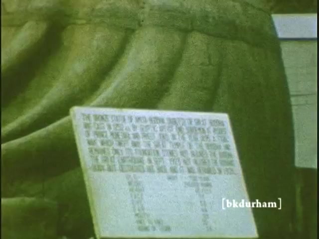 The still captures the Great Buddha (Daibutsu) in Kamakura, Japan, taken from a home movie in 1959/1960. Prominently displayed in the foreground is a sign with text, likely providing information about the statue. The Buddha's serene features and the texture of its robes are partially visible, showcasing the craftsmanship. The muted colors and graininess typical of 16mm film evoke a nostalgic atmosphere.