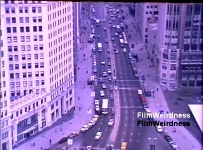 The still shows a busy urban street scene from an elevated view, likely in a city environment. Various vehicles, including cars and trucks, are moving along the divided road, while numerous pedestrians occupy the sidewalks. The architecture is diverse, featuring large buildings that suggest a commercial area. The color tone appears slightly faded, characteristic of 16mm film from the 1980s. The image captures a moment in time, emphasizing commerce and activity during that era.