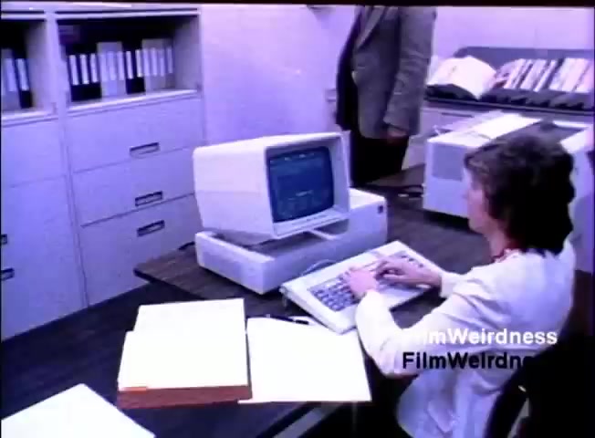 The still depicts a woman typing on a keyboard connected to an IBM System 36 computer, which features a monitor displaying text-based information. She is seated at a desk cluttered with documents and papers. In the background, a man stands near filing cabinets filled with labeled folders, suggesting a business environment. The overall setting reflects the 1980s office culture, emphasizing the use of technology in business processes.