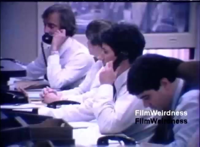 The still depicts a group of four men in an office setting, all engaged in phone conversations. They are dressed in formal attire, featuring shirts and ties, with a focus on a professional atmosphere. The background suggests an office environment, possibly with desks and equipment typical of the 1980s. The scene conveys a sense of busy corporate activity, likely related to sales or customer service for the IBM System 36.