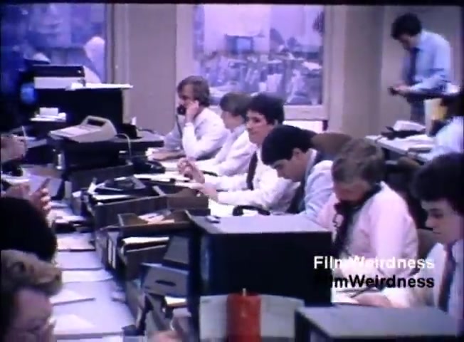 The still shows an office environment, likely from the 1980s, featuring several young men in white dress shirts seated at desks. Some are engaged in phone conversations, while others are focused on paperwork. The atmosphere appears busy and industrious, with stacks of papers and office equipment visible. The lighting has a muted, slightly purple hue, suggesting the film's age. This scene illustrates the corporate culture of the time, emphasizing communication and work in sales.