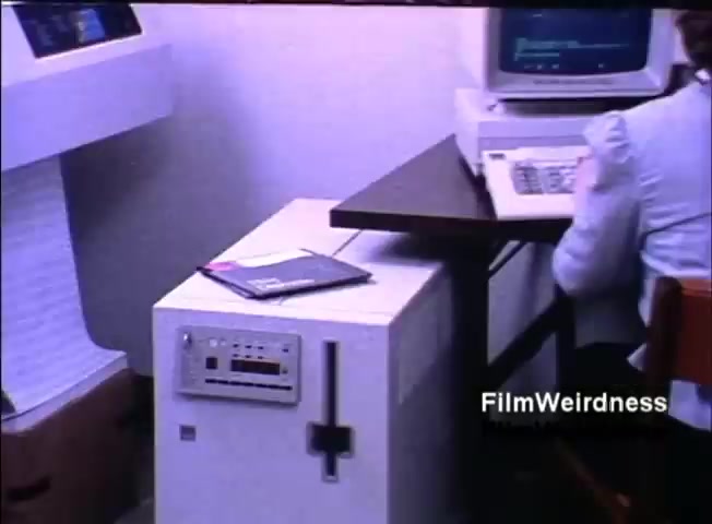 The still features a vintage office setup from 1984, showcasing an IBM System 36 computer on the left. The unit is boxy and white, with a control panel visible on the front. Next to it, there’s a small table supporting a computer monitor and a keyboard, indicating a user is engaged with the system. A notebook lies on the table, contributing to the typical office environment of that era. The overall atmosphere reflects technological advancement during the early 1980s.