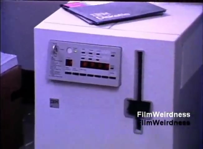 The still depicts an IBM System 36, a computer from the 1980s, featuring a prominent front panel with buttons and a digital display. It's a white machine, showcasing its utilitarian design, and there's a brochure or manual resting on top. The setting suggests a business environment typical of that era.