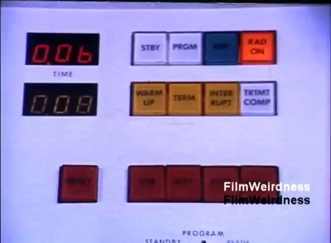The still features a control panel from an IBM System 36, showcasing various buttons and displays. It includes digital readouts labeled 'TIME' and 'RAD ON,' with indicator lights. The buttons are color-coded, featuring functions such as 'STBY,' 'PRGM,' and 'WARM UP.' The layout is functional, typical of 1980s computer hardware.
