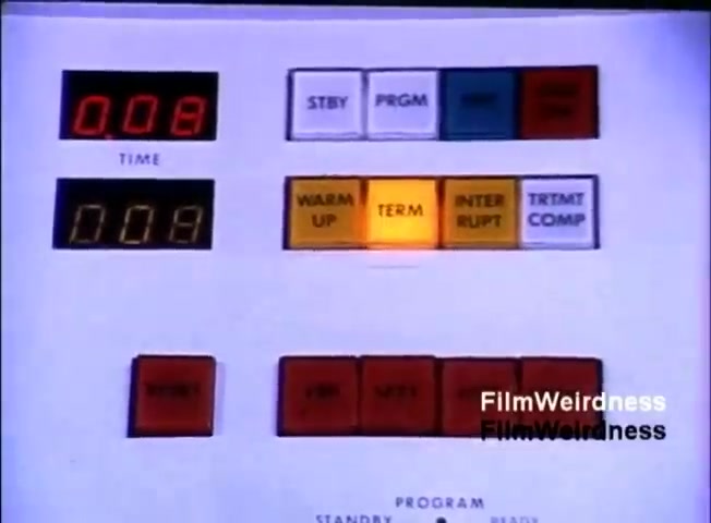 The still features a control panel from the IBM System 36, displaying a digital timer reading '0.08' and various colored buttons. Notable buttons include 'WARM UP,' 'TERM,' and 'INTER RUPT,' with 'TERM' illuminated. It emphasizes the technological interface of the era, reflecting the design and functionality of computing systems in 1984.