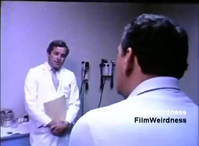 The still depicts a scene from the IBM System 36 sales film, showcasing two male characters in a medical setting. One man, dressed in a white lab coat with a clipboard, appears to be speaking with another man, who is wearing a white shirt and facing away from the camera. The environment suggests a doctor's office, indicated by the presence of medical equipment in the background. The overall tone seems professional, likely relating to a discussion about medical technology or systems relevant to IBM's offerings at the time.