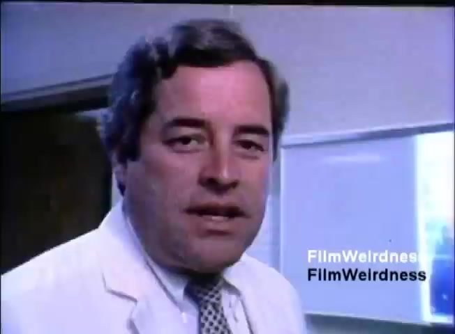 The still features a man wearing a white coat and a patterned tie, standing in an office environment. He has short, styled hair and is looking directly at the camera, likely speaking or presenting. The background is slightly blurred but suggests a professional setting, possibly with a whiteboard or glass partition visible. The ambiance is typical of corporate training or sales materials from the 1980s.