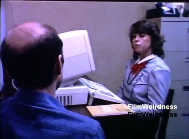 The still depicts a scene from the IBM System 36 sales film from 1984. It features a man with a receding hairline seated at a desk, facing a woman dressed in a light blue blazer. She appears to be engaged in conversation while sitting in front of a computer monitor and a stack of documents. The setting suggests a business or office environment, likely focused on a sales pitch or discussion about the IBM System 36.