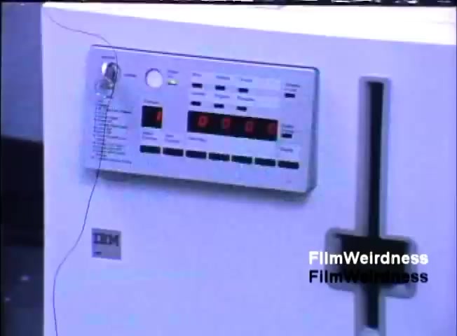 The still features a white device, likely a computer or terminal, with a prominent control panel that includes buttons and a digital display. The IBM logo is visible, indicating the brand. Wires are attached to the device, suggesting it may be connected to additional equipment or designed for specific functionality. The aesthetic reflects the technology of the early 1980s, portraying a vintage computing environment.