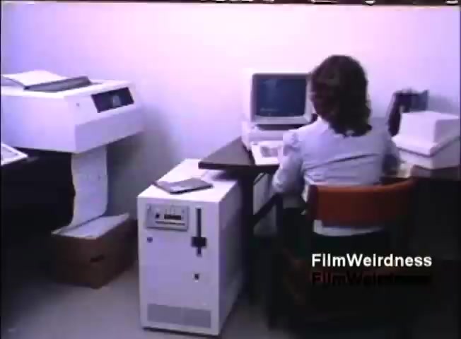 The still features an office setting typical of the 1980s, showcasing a woman sitting at a desk in front of a computer monitor. Next to her, there's a white printer or copier, and a small, square white device that appears to be a computer system, possibly the IBM System 36. The environment is simple and functional, with minimal furnishings and a professional ambiance.