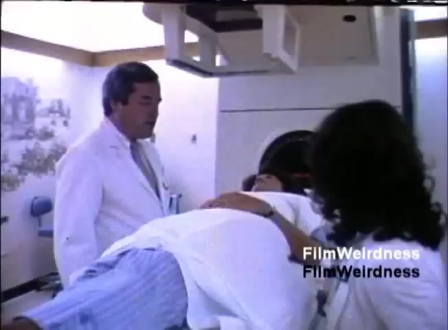 The still features a medical setting where a patient lies on an examination table, attended by two individuals in white lab coats—a man and a woman. The man appears to be explaining something to the patient, while the woman assists. The environment is clinical, with medical equipment visible in the background, suggesting a focus on healthcare technology, likely related to the sales pitch for the IBM System 36 system in a medical context.