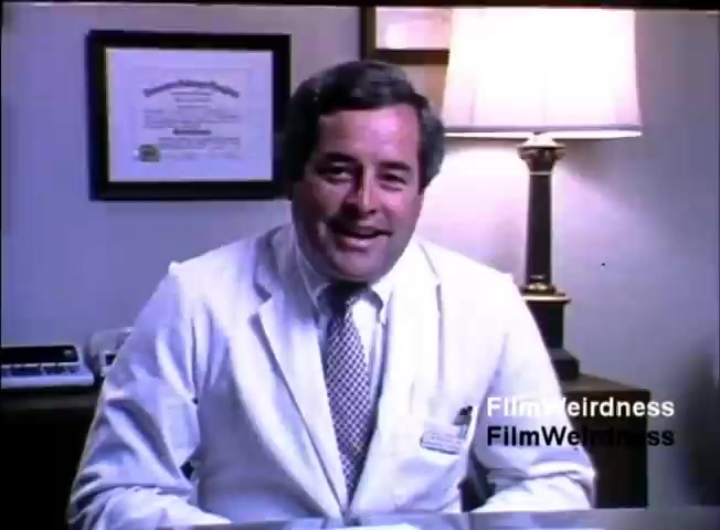 The still features a smiling man in a white lab coat, seated at a desk in an office setting. Behind him are framed certificates and a lamp, indicating a professional environment. The man appears to be engaging, likely promoting or discussing the IBM System 36, with a focus on its benefits or applications. The setting reflects a blend of healthcare and technology, characteristic of the era's corporate promotional materials.