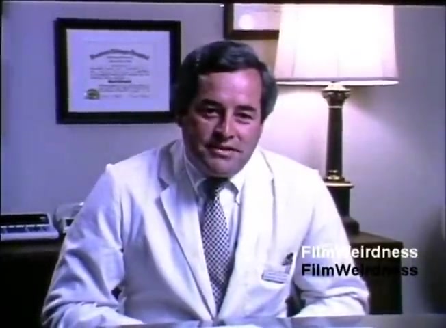 The still features a man wearing a white lab coat and a patterned tie, seated at a desk. Behind him, there's a lamp and a framed document or certificate on the wall. The setting appears to be an office, likely for a medical or professional context. The overall tone is formal, and the man presents a friendly demeanor, likely addressing an audience as part of the IBM System 36 sales film from 1984.
