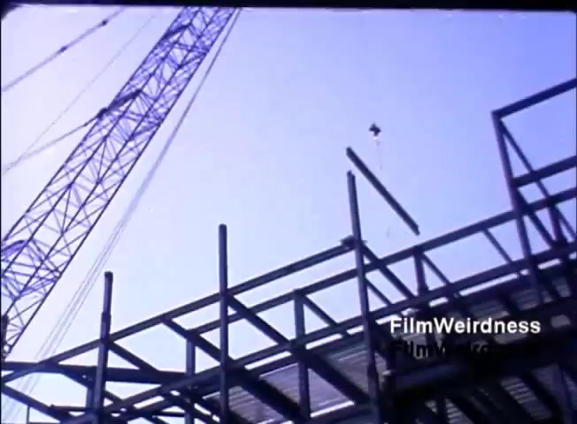 The still shows a construction site, featuring a crane lifting a long beam towards a steel framework. The sky is clear, contributing to a bright atmosphere. The focus on construction suggests themes of innovation and progress, aligning with the promotional intent of the IBM System 36 sales film from 1984.